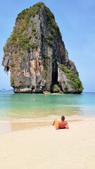 Wall Mural - A man is relaxing on the sandy beach with a towering rock formation in the background. natural landscape includes trees, a tranquil body of water, and a clear sky above Railay Beach, Krabi, Thailand