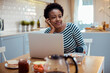 © Davor - Black woman enjoying breakfast while working on laptop in a sunny kitchen