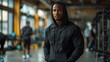 A serious muscular African-American male instructor stands with his hands crammed into a hoodie and a hood over his head in a modern fitness center and looks into the camera