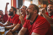 A group of men in red sports jerseys are sitting at the bar laughing and cheering for their team, watching football on TV together with friends while drinking beer