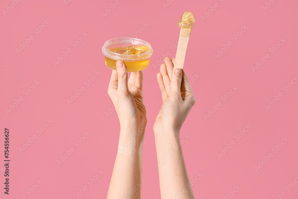 Female hands holding container with sugaring paste and spatula on pink background