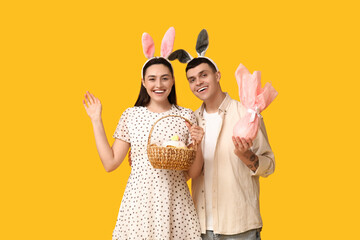 Poster - Happy young couple with Easter bunny ears, basket and gift on yellow background