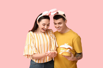 Poster - Happy young couple with Easter cake, eggs and bunny ears on pink background