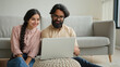 © Yuliia - Multiracial couple Arabian man and Indian woman on floor family couple homeowners wife girlfriend and husband boyfriend watching laptop movie smart TV enjoy home leisure smiling talk video call smile