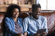 Laughing African couple sit on sofa with digital tablet, feels overjoyed have fun watch comical videos funny online content, joking, looking carefree resting at home. Modern tech usage, humor concept
