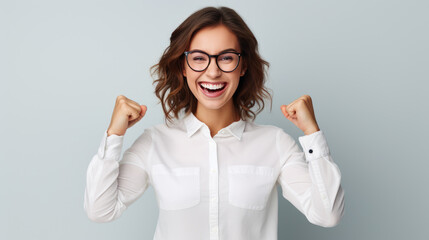 Poster - Joyous woman with curly hair and glasses, raising her fists in a triumphant gesture, wearing a white shirt, and smiling against grey background