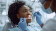 © Fokke Baarssen - African American female doctor in mask and gloves doing the dental procedure with mirror and metal explorer for the patient lying under a lamp in the medical room, Modern medical equipment dentist