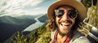 A man with a hat and sunglasses is seen smiling while taking a selfie in the mountains. The sunny weather highlights his casual attire as he captures the picturesque landscape behind him.