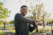 Young black man in sports clothing smiling while doing stretches at park