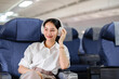 © Nathamanee - Asian woman sitting and using a smartphone in an airplane Listening to music next to the airplane seat window travel and technology concept