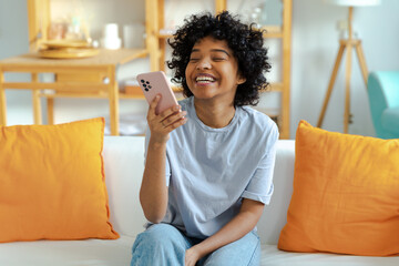 Wall Mural - Excited happy young black african american woman holding cell phone laughing feeling joy getting mobile message. Overjoyed girl laughing aloud sitting on couch watching funny video reading news