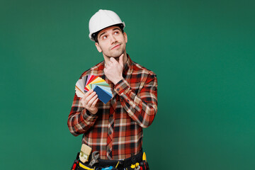 Sticker - Young minded laborer man wear red shirt hardhat hat hold color wallpaper palette look aside isolated on plain green background. Instruments accessories, renovation apartment room. Repair home concept.