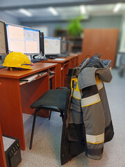 The engineer's yellow work clothes and helmet hang on the back of a chair in the office near the computer. The workplace of a specialist.