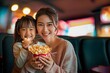 A woman and a young girl smiling and sharing a popcorn at a cinema