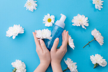 Wall Mural - Concept natural cosmetics for facial skin care. Woman hands with bottle with water foam for cleansing on blue background with white flowers, top view