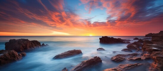 Poster - A fluid sunset over the ocean with rocks in the foreground creating a stunning natural landscape art, blending water, sky, and clouds on the horizon