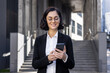 © Tetiana - Close-up photo of a smiling young business woman in a suit and glasses standing outside an office building and using a mobile phone