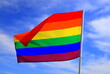Realistic rainbow flag of LGBT organization waving against a blue sky. LGBT pride flags include lesbians, gays, bisexuals and transgender people.