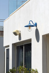 a white building exterior with two distinct light fixtures, modern security light, and a classic blue sconce with bulb. Both are mounted above windows casting shadows on the wall in bright sunlight