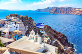 Fototapeta Tulipany - Greek orthodox church with bells, greek flag and famous white houses on Santorini island, Aegean sea, Greece.