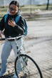 © qunica.com - Casual outdoor shot of a cheerful young woman with glasses, using a smart phone while leaning on a bicycle with a backpack.