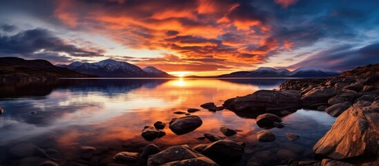 Poster - A picturesque sunset over a tranquil lake, with rocks in the foreground, mountains in the background, and a colorful sky filled with clouds and an afterglow