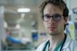 A young male doctor with unkempt hair wearing eyeglasses and a white lab coat in a hospital looking at the camera.