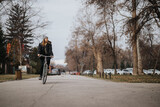 Fototapeta  - An adult female cyclist rides leisurely on a park path, surrounded by autumn trees and a calm urban environment, embodying active lifestyle and serenity.