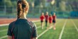 © tashechka - A focused young female athlete standing on a track field, with teammates in the distance.