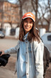 A happy young dark-haired woman in a cap and a denim jacket walking on a spring day in the city.