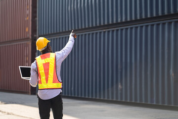 African American logistic engineer man worker or foreman holding notebook computer with finger point at container site	