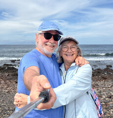 Sticker - Carefree senior caucasian couple hugging standing outdoors together at the beach takes photos with selfie stick enjoying vacation and retirement at sea