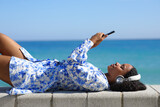 Fototapeta Pomosty - Happy black woman watching media lying on the beach