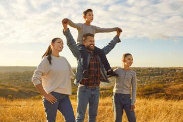 Portrait of happy joyful family standing in the field with two kids and looking into the distance with hands up enjoying beautiful nature together. Young parents with children walking outdoors.