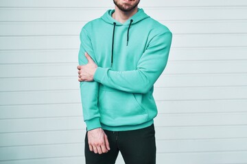 Canvas Print - A Man in a Stylish Blue Hoodie Leans Against a White Wall. Logo and brand swag mock-up. Isolated horizontal photo.