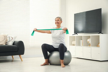 Wall Mural - Senior woman doing exercise with elastic resistance band on fitness ball at home