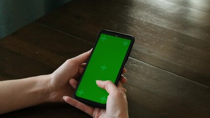 Wall Mural - A woman use a phone with a green screen while sitting at a table, viewed from over her shoulder. Scrolling. Video footage