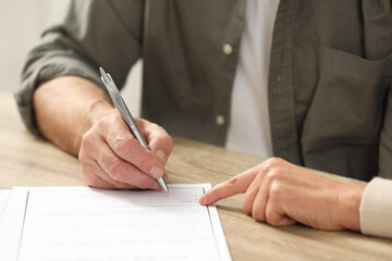 Notary pointing to senior man where to sign Last Will and Testament at wooden table indoors, closeup