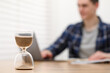 © New Africa - Hourglass with flowing sand on desk. Man taking notes while using laptop indoors, selective focus