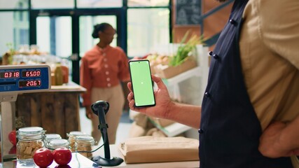 Young man holds smartphone at store checkout, presenting isolated greenscreen display on mobile device. Farmer and vendor using blank chromakey template with copyspace technology.