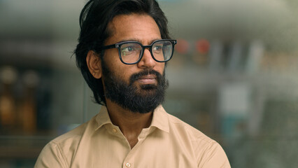 Medium portrait handsome Indian entrepreneur in glasses in creative office look to side thoughtful thinking pondering Arabian man businessman contemplating e-business ideas business executive think