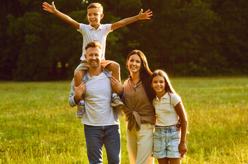 Wall Mural - Happy young smiling family of four with son and daughter standing in the summer park and looking cheerful at camera. Mother, father with two kids walking in nature enjoying sunny day together.