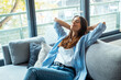 © simona - Front view portrait of a relaxed person smiling sitting on a couch in the living room at home with a window and a city in background. One woman relaxing indoor on the couch alone with happy expression