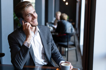 Wall Mural - Cheerful businessman dressed in formal wear sitting in cafe and look aside while talking by phone and using laptop.