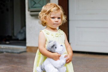 Wall Mural - Little toddler girl playing with soft toy dog. Cute baby child holding toy and plays outdoors.