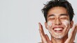 Portrait of a Asian man his cheeks covered in cream, smiles while covering his eyes during a skincare routine against clean background with space for text, Generative AI.