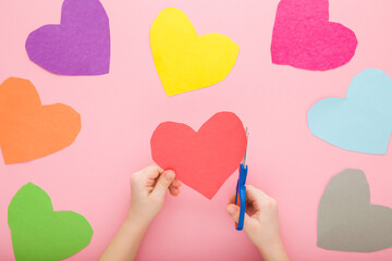 Wall Mural - Little child hands holding scissors and cutting colorful heart shapes from application paper on pastel pink table background. Making decoration elements. Closeup. Point of view shot. Top down view.