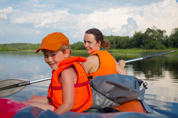 Little boy and woman in life-jackets sail on inflatable boat on river at summer, focus on woman