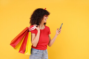 Wall Mural - Happy young woman with shopping bags and modern smartphone on yellow background