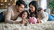 © MP Studio - Happy family consisting of a mother, father, and young daughter lying on a rug at home, smiling at each other with a piggy bank in the foreground.